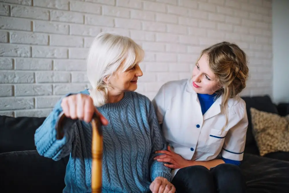 Two people sitting on a couch, one holding a cane. One person wears a blue sweater, the other in a white uniform.