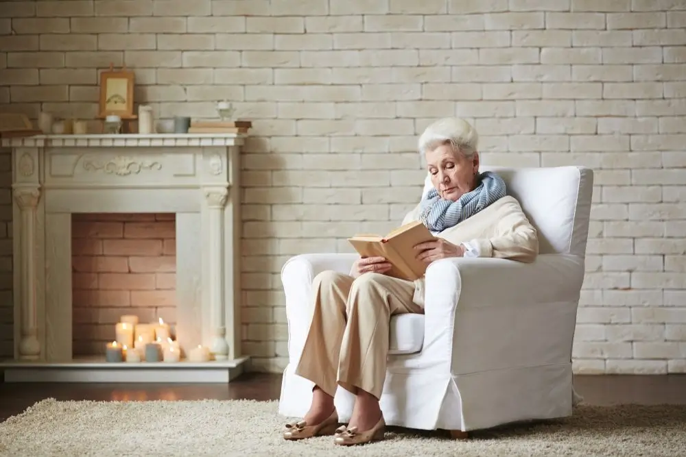 A person reads on a white armchair in a cozy room with candles on a decorative fireplace and a textured brick wall.
