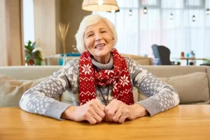 A person wearing a festive red scarf and gray sweater sits smiling at a wooden table in a cozy, well-lit room.